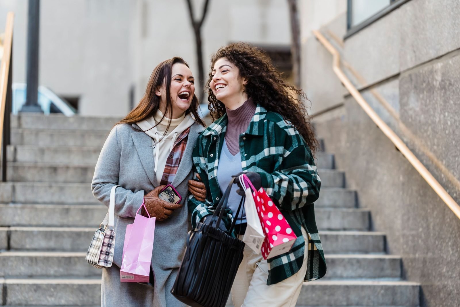 Two friends laughing together on city steps, carrying shopping bags