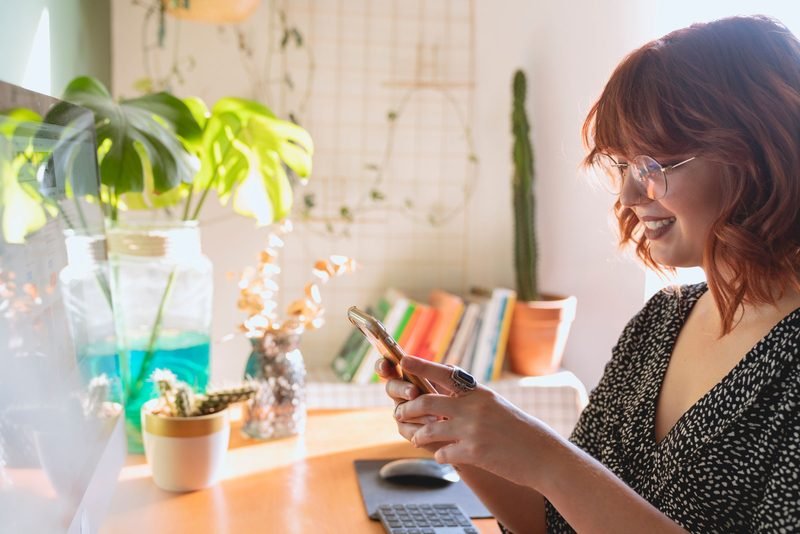 Smiling woman using her phone at a sunny desk with plants